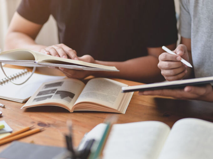 Partial view of hands and arms of two people at a work surface with open books, a tablet, stylus, sticky notes, pencils, etc.