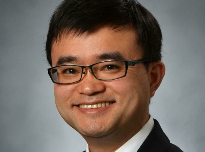 A man with neatly-trimmed brown hair and glasses smiles in a dark jacket, white shirt, and tie.